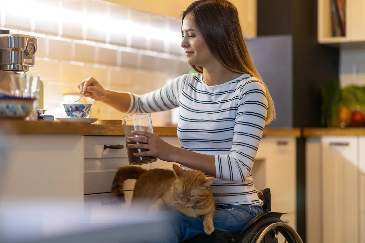 A woman in a wheelchair in the kitchen, preparing a cup of tea. An orange cat is on her lap.