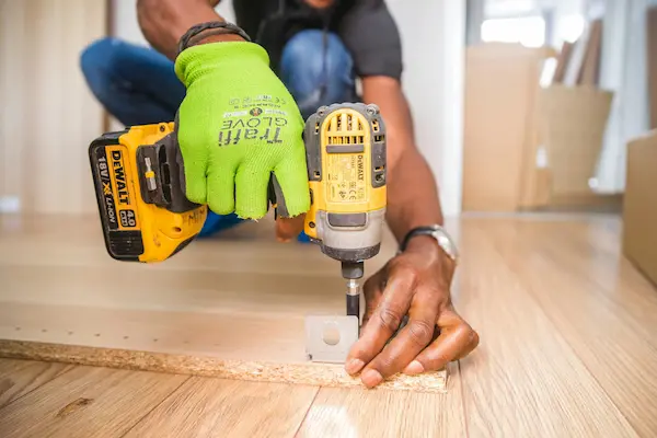 A man uses a drill to screw in a bracket to a piece of wood on the floor of a living room.