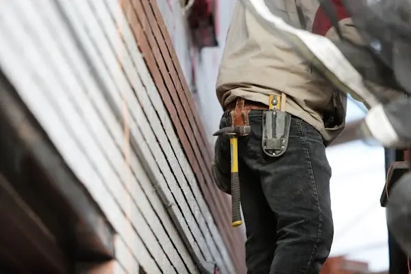 A man in denim stands outside the side of a building with a tool belt wrapped around his waist.