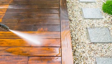 A pressure washer blasting a jet of water at a wooden deck while a handyman provides pressure washing service to clean away a layer of grime on the deck.