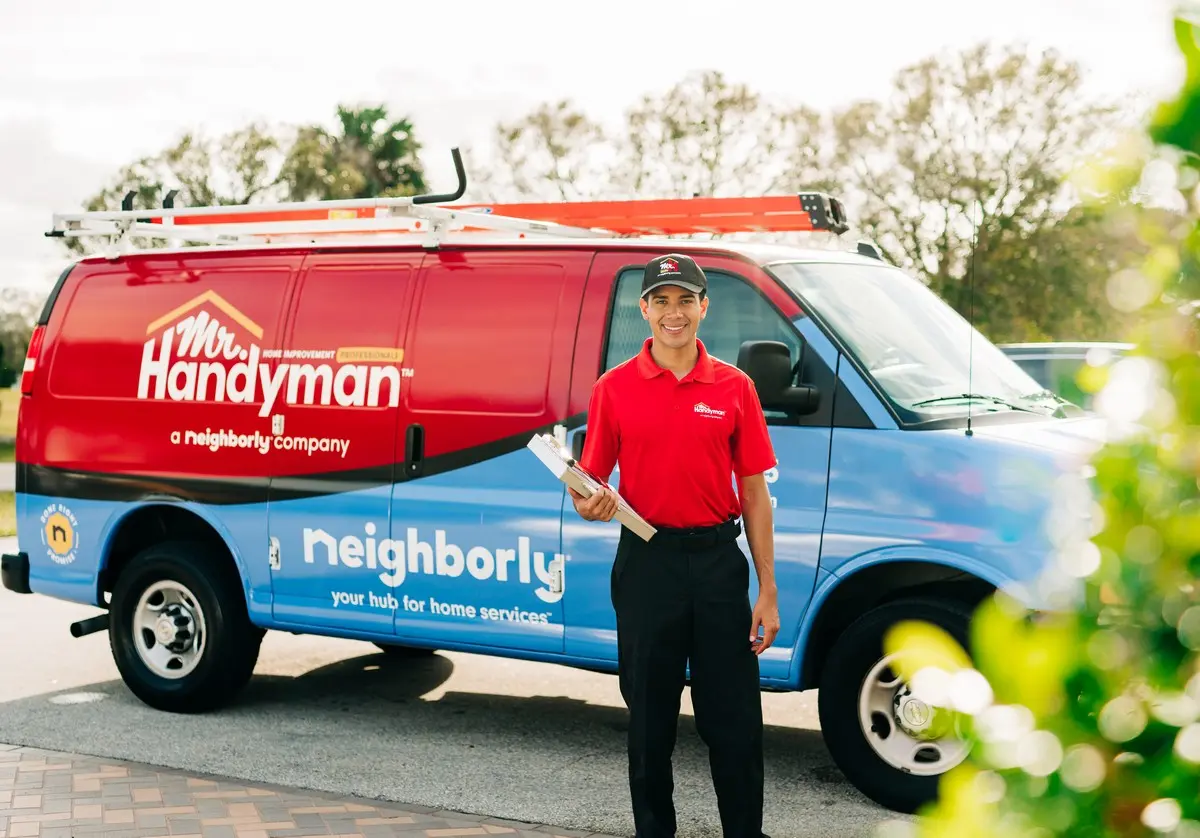 A Mr. Handyman technician standing in front of a company van holding a clipboard.