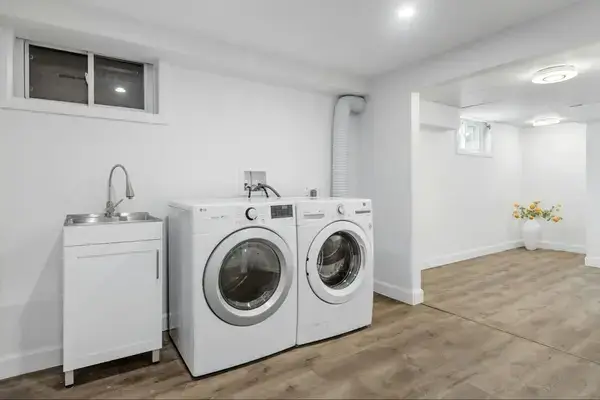 A white sink, washer, and dryer sit in a brightly lit room with white walls and wooden floors.