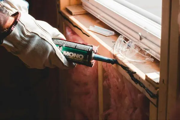 A pair of gloved hands are holding a tube of sealant and applying it to a window pane.