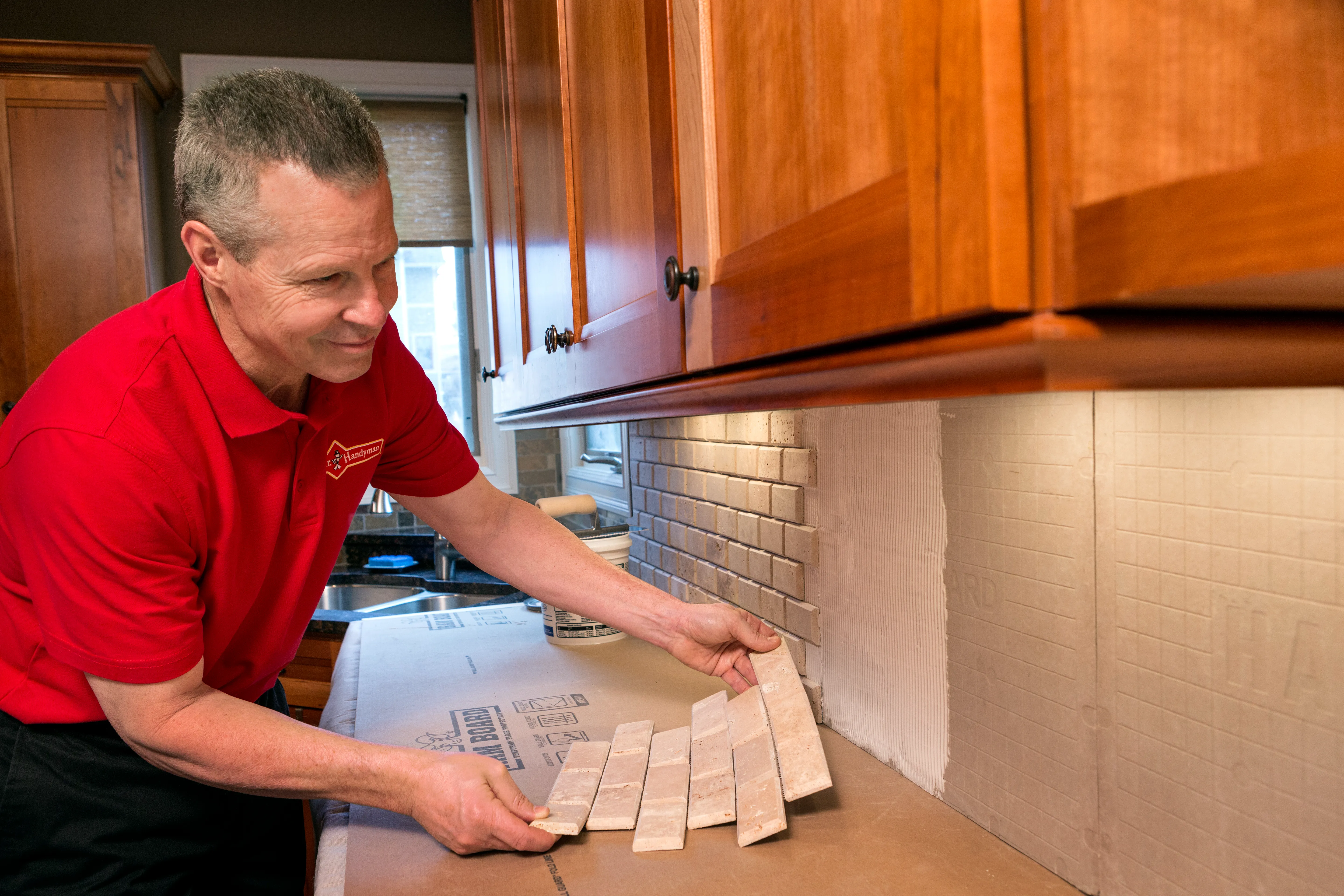A handyman from Mr. Handyman installing a kitchen backsplash by mounting large sections of tile.